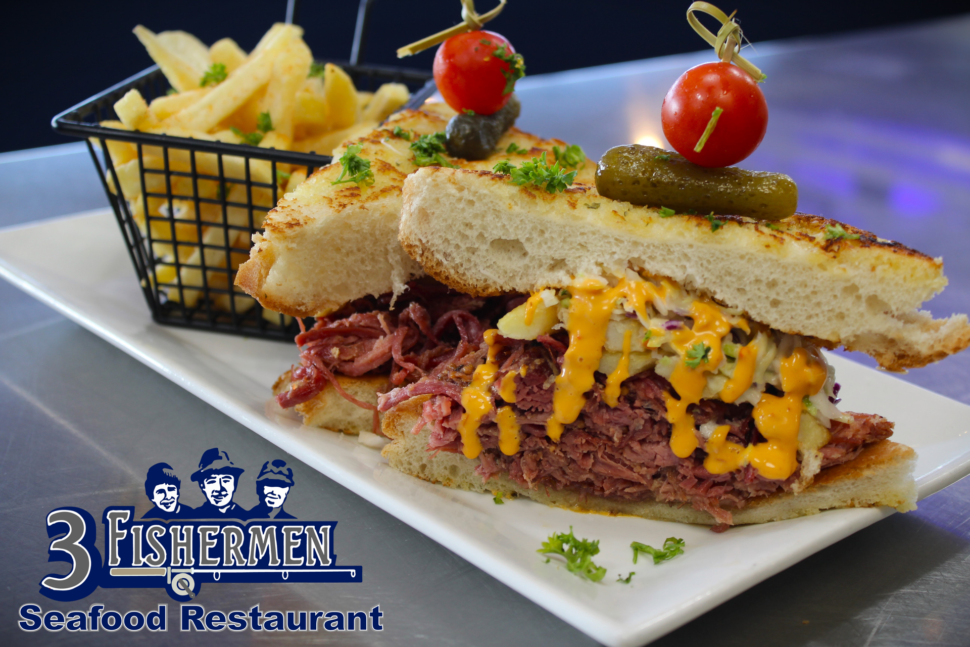 A corned beef sandwich is on a white plate next to a basket of french fries