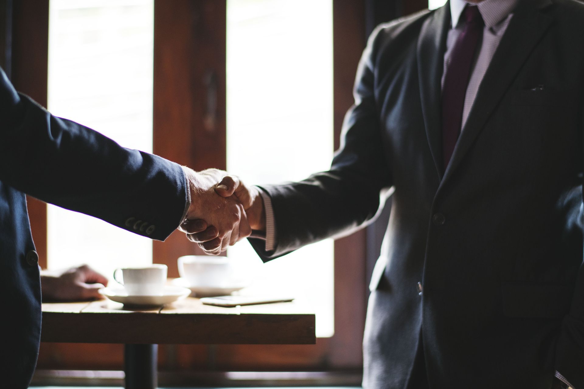 Two men in suits are shaking hands over a table.