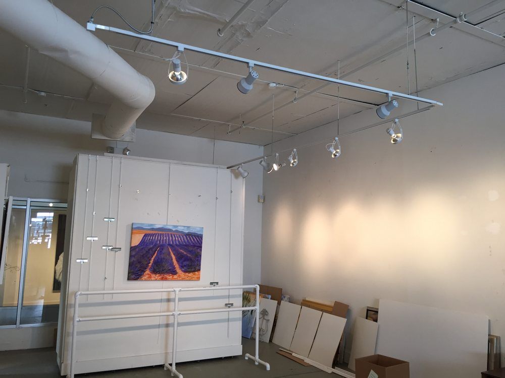 Empty gallery space with track lighting, white walls, and artwork.