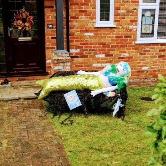A mermaid scarecrow on a small stone structure in a front yard next to a brick house.