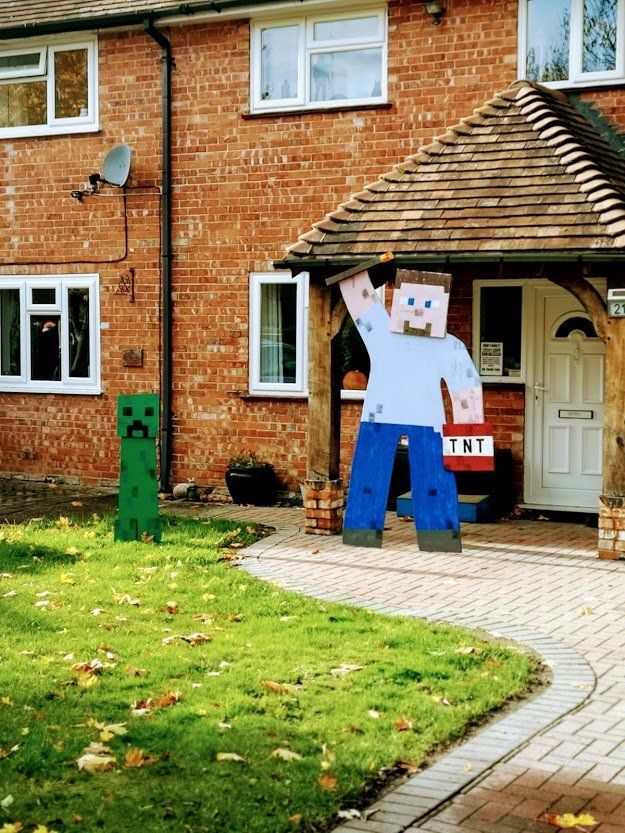 A Minecraft-themed yard display with a Steve figure waving, a creeper, and a TNT sign in front of a brick house.