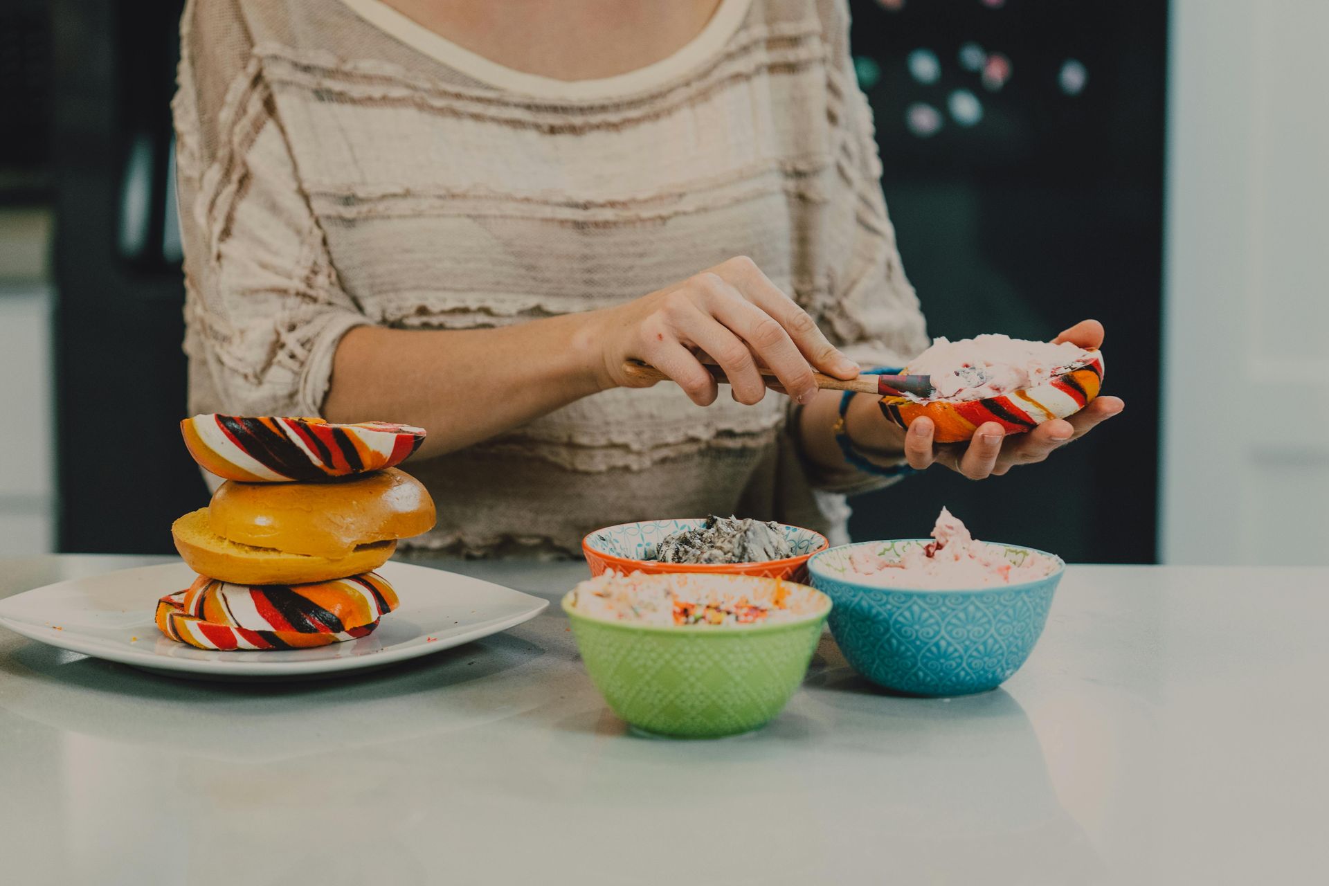 Woman decorating a glazed donut with sprinkles; bowls of toppings, stacked donuts on a plate.