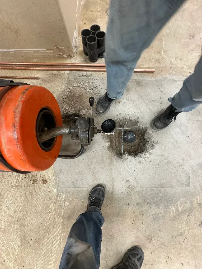 A drain cleaning machine is positioned over a hole in a concrete floor, with two people standing nearby.