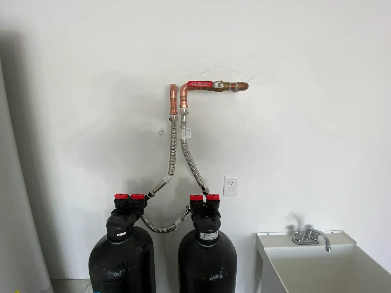 Two black water filtration tanks connected by braided metal hoses to copper wall pipes above a utility sink.