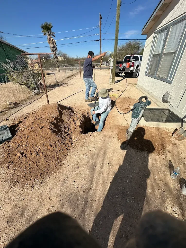 Two people use a jackhammer to excavate a trench next to a house on a sunny day.