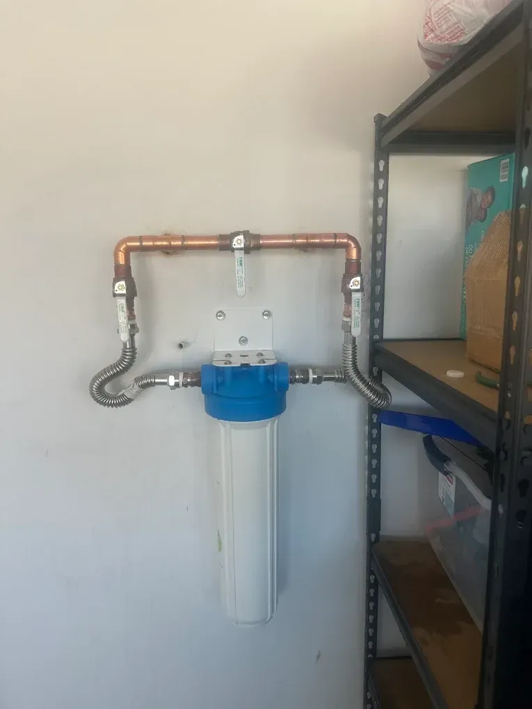 A water filtration system mounted on a wall with copper piping, a blue filter housing, and a bypass valve handle.