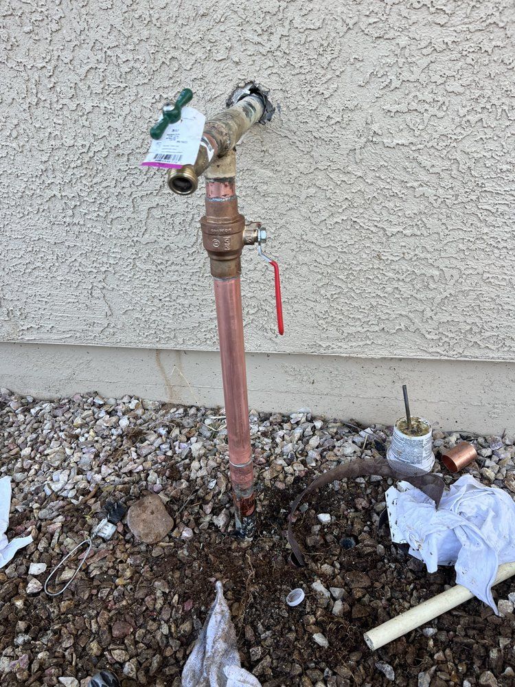 A copper pipe and hose bib assembly installed against a beige stucco wall, with construction debris on the ground below.