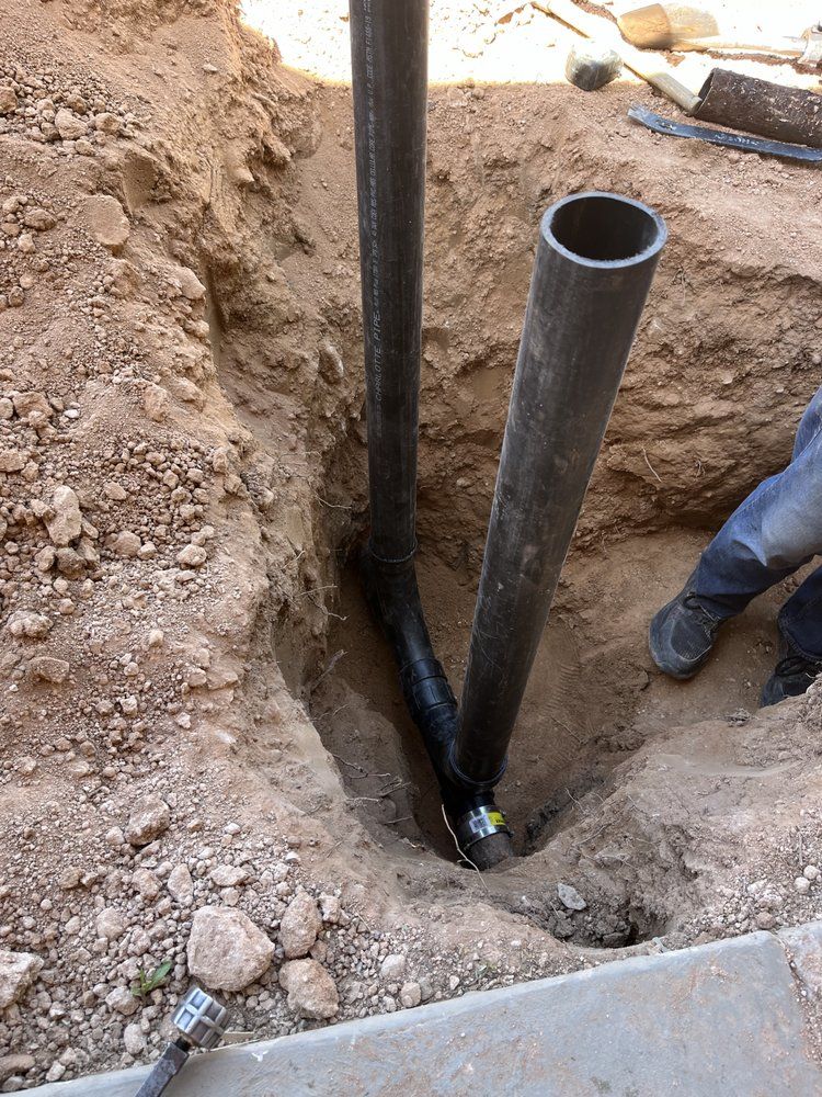 Two dark pipes meet at a T-fitting inside a dirt trench, with a person's legs visible on the right side of the hole.