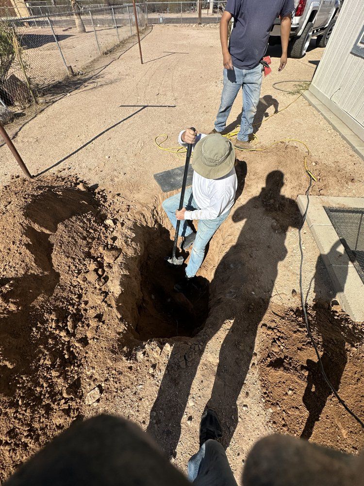 A person in a sun hat digs a hole in a dirt yard while another person stands nearby.