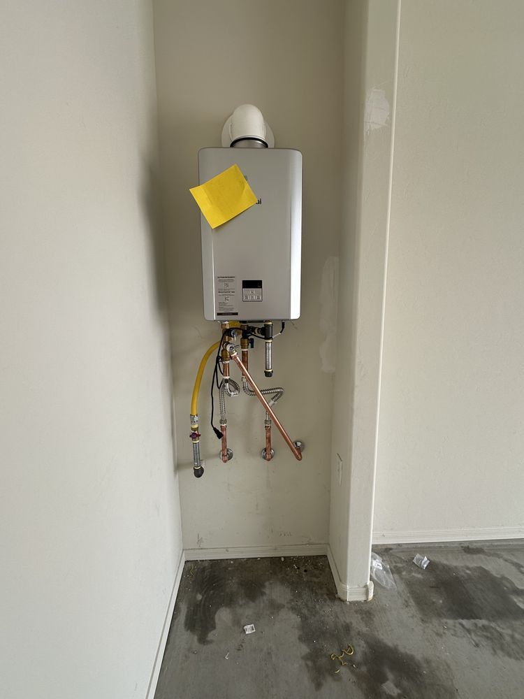 A silver tankless water heater with a yellow note attached, mounted on a white wall above exposed pipes in a garage.