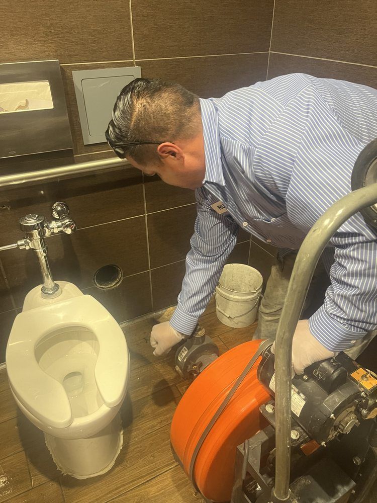A professional plumber uses a drain snake machine to clear a clogged toilet in a tiled restroom.
