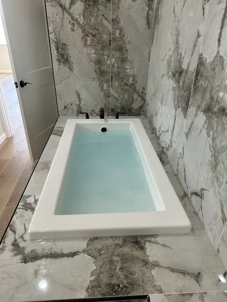 A white rectangular bathtub filled with water, surrounded by gray and white marble tile on the walls and deck.