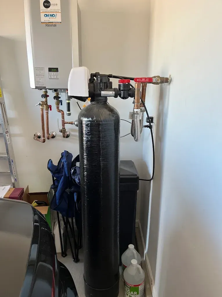 A black water filtration tank stands in a garage next to a wall-mounted water heater and plumbing connections.