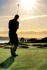 Golfer silhouetted on a green course at sunset, holding a club after a swing.