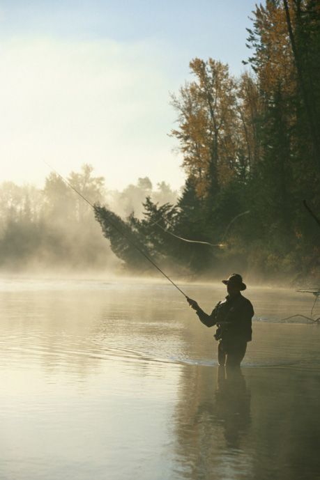 Person fishing in a misty river at sunrise, with trees reflected in the calm water.