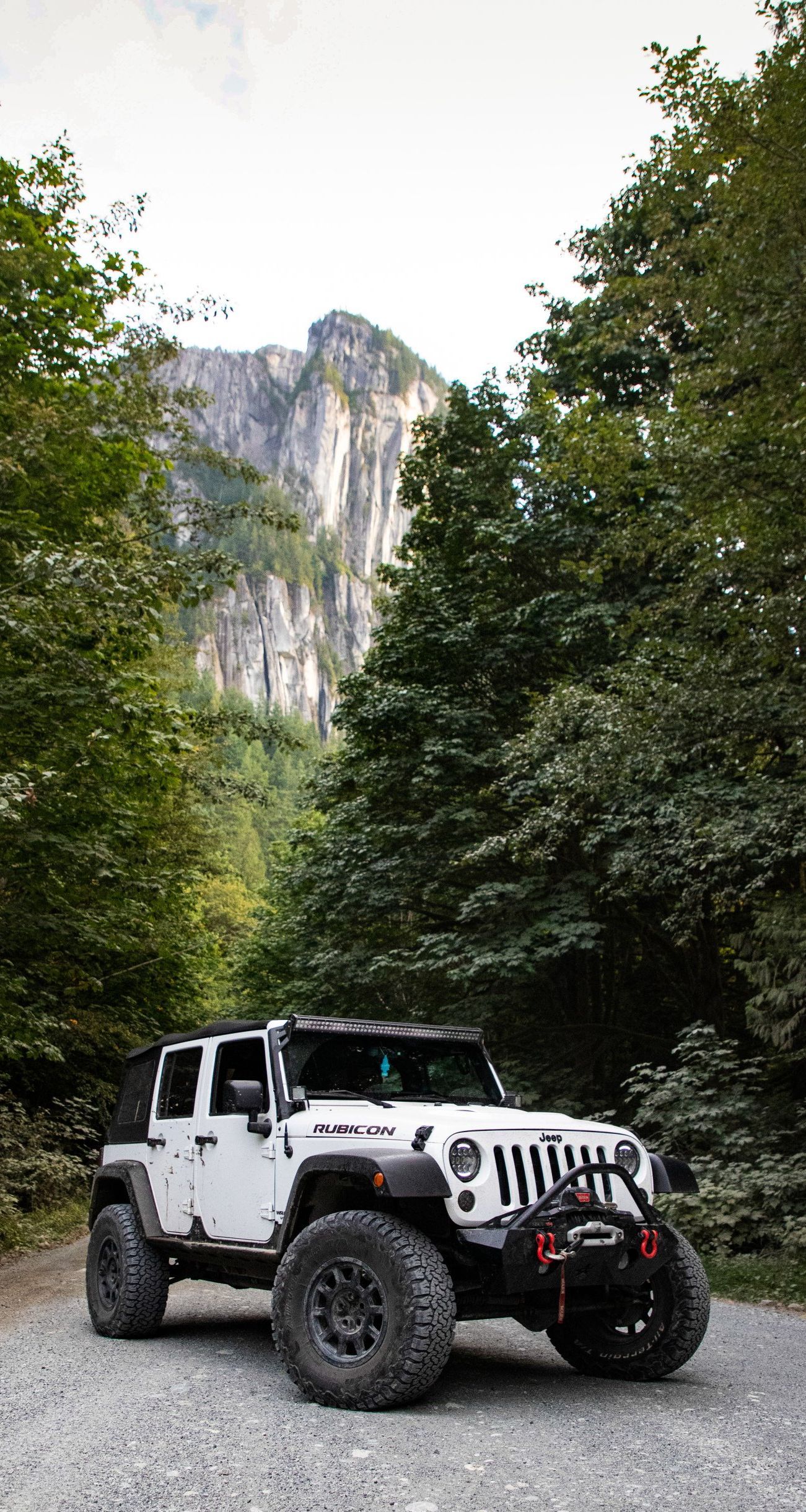 White Jeep parked on a gravel road in a forest with a large rock face in the background.