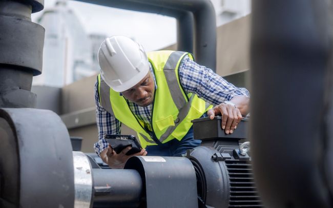Technician in hard hat and safety vest inspects industrial machinery with a handheld device.