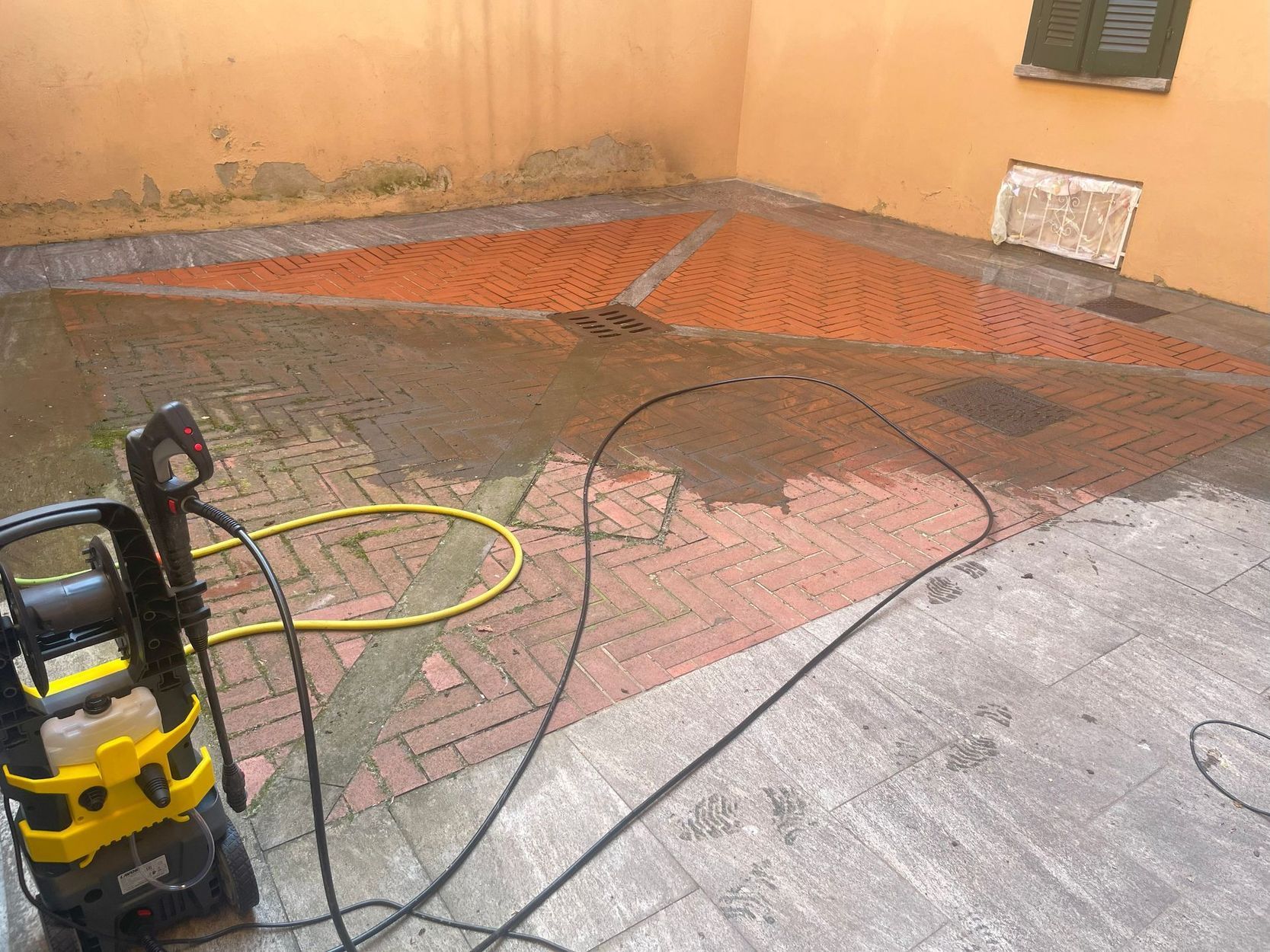 Pulizia di un patio in cemento bagnato con un’idropulitrice, accanto a un muro arancione e a una recinzione nera.