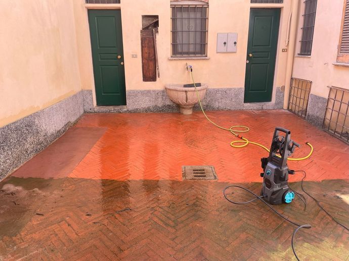 Piccolo cortile con pavimento arancione, due porte verdi, una fontana a muro e un’idropulitrice con tubo.