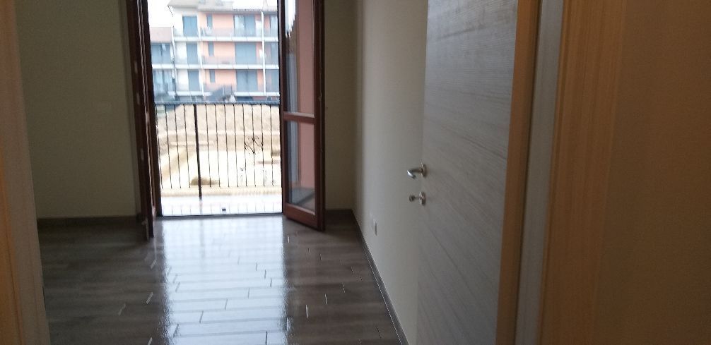 Corridoio vuoto con pavimenti scuri, pareti bianche e una porta aperta su un balcone o un patio.