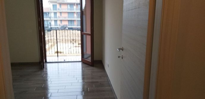 Corridoio vuoto con pavimenti scuri, pareti bianche e una porta aperta su un balcone o un patio.