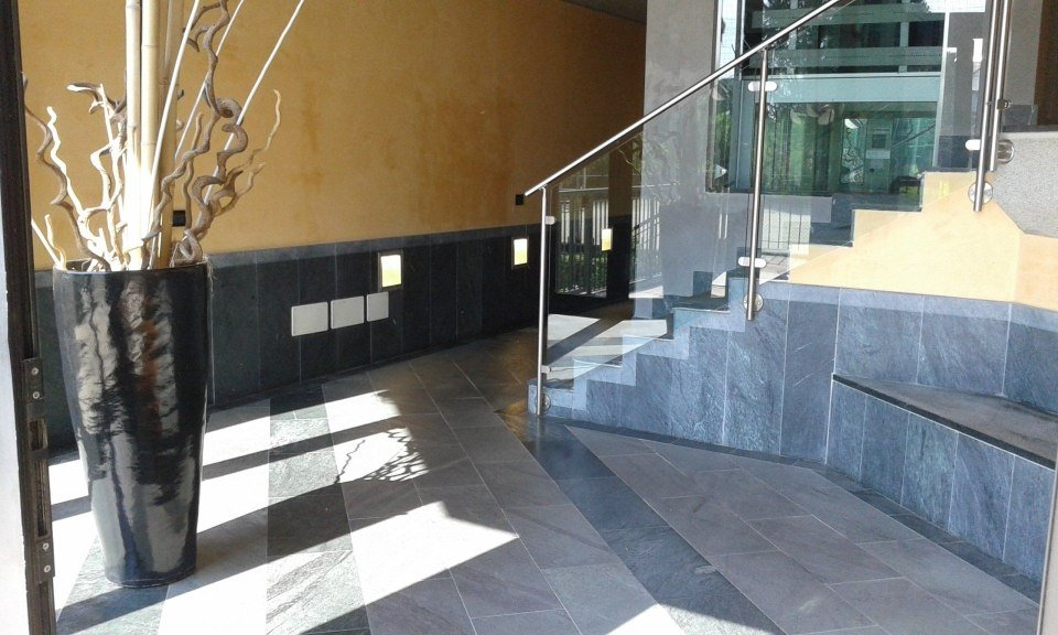 Atrio moderno con pavimento piastrellato, fioriera nera, scala e parapetto in vetro accanto a una parete illuminata dal sole