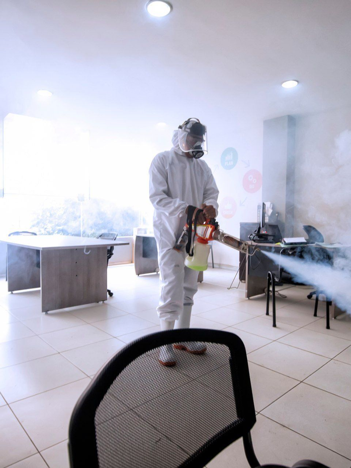 Una persona con indumenti protettivi spruzza un disinfettante in una stanza luminosa con sedie e un tavolo, mentre la nebulizzazione si diffonde nell’aria