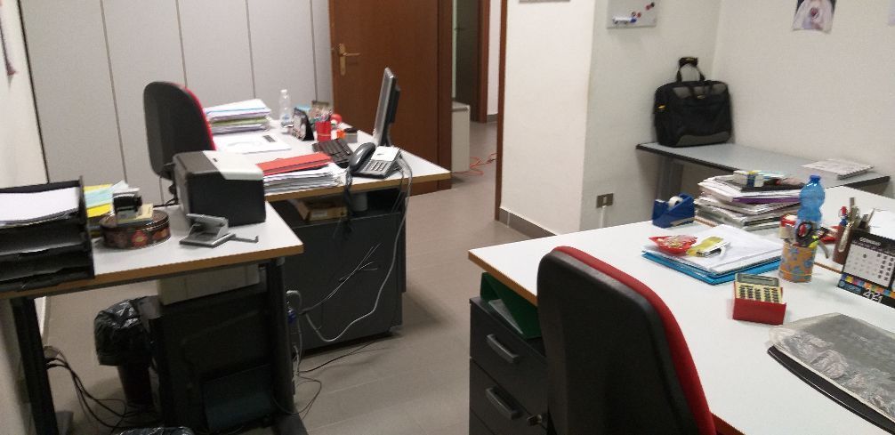 Ufficio disordinato con scrivanie, monitor, sedia, fogli e materiale da ufficio in una stanza piccola.