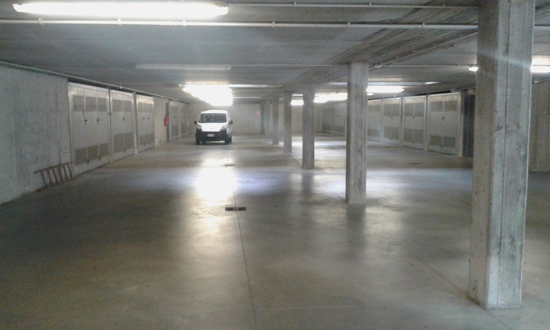 Parcheggio coperto in cemento vuoto, con pilastri portanti e un furgone bianco in lontananza.