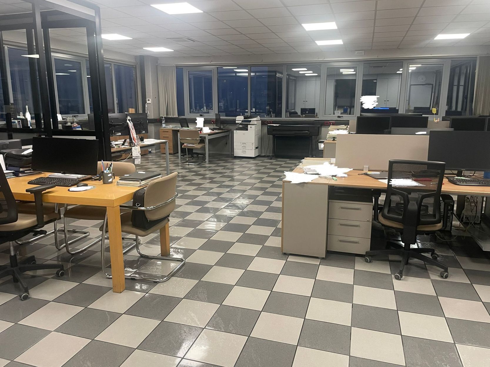 Ufficio vuoto con pavimento a scacchi, scrivanie, sedie e postazioni computer sotto luci intense a soffitto