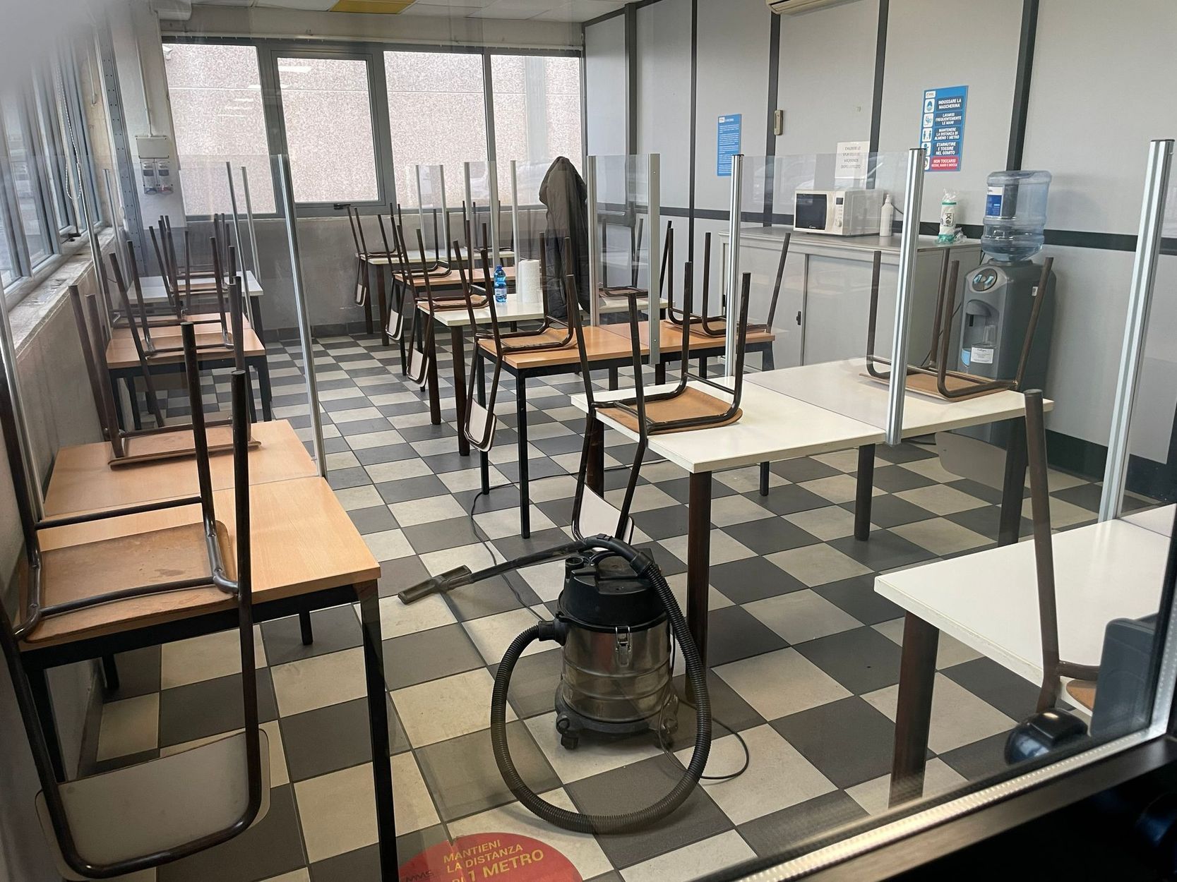 Aula vuota con i banchi distanziati e un ventilatore a terra su un pavimento a scacchi bianchi e neri