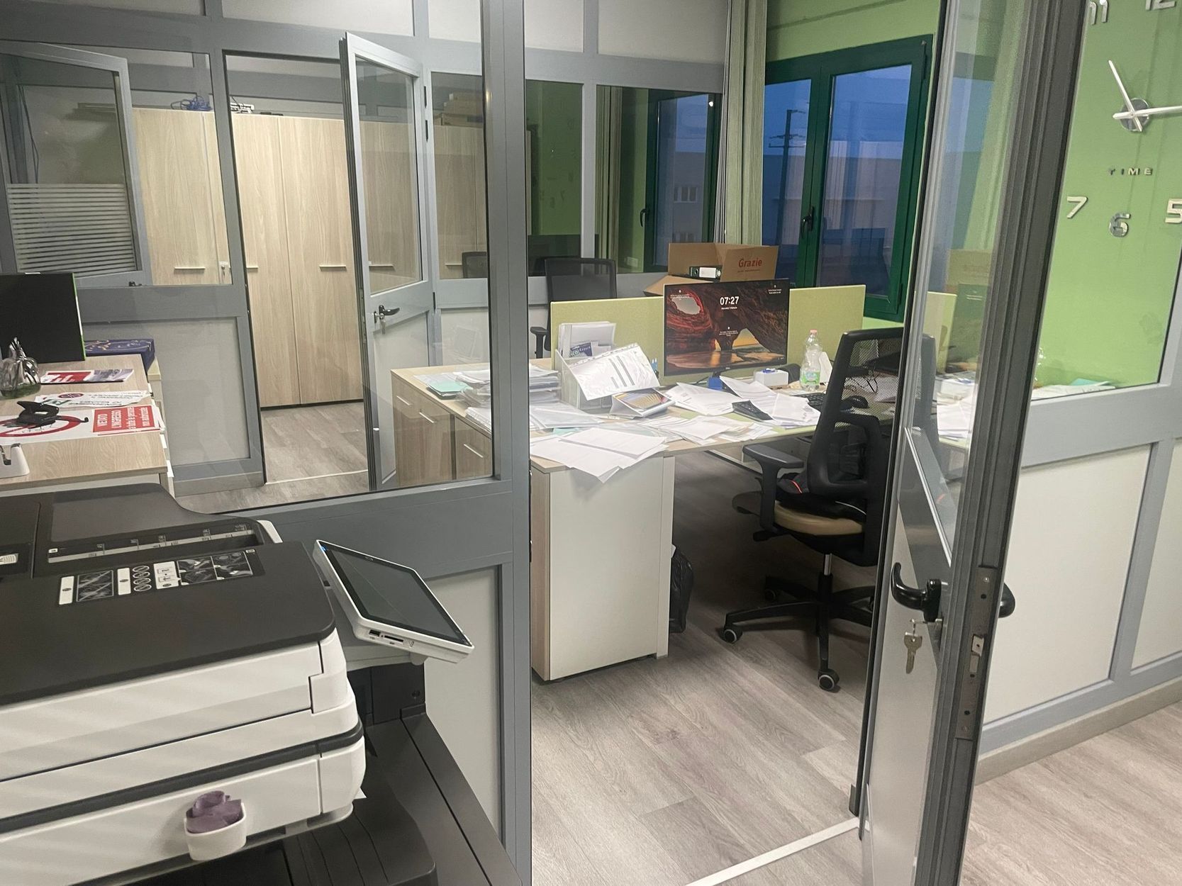 Disordinato cubicolo da ufficio con scrivania, sedia, fogli e schedari dietro pareti di vetro