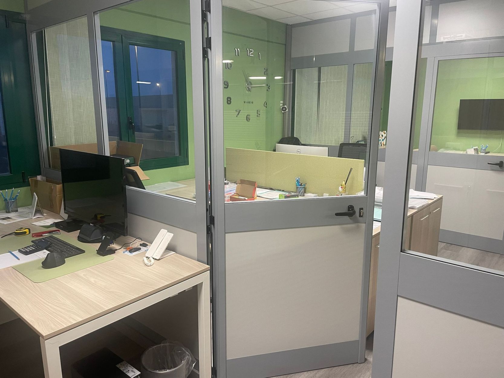 Postazioni d’ufficio vuote, con scrivanie, computer e pareti verdi, in un ambiente suddiviso da pareti divisorie.
