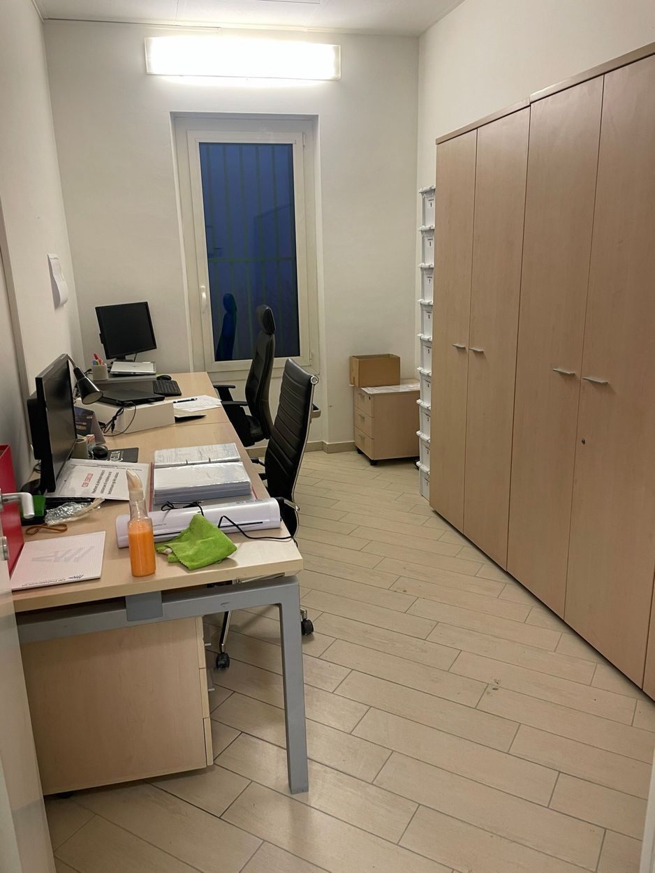 Piccolo ufficio con scrivanie, computer, documenti e alti armadi in legno addossati alla parete.