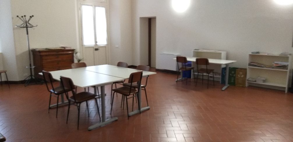 Stanza vuota con tavoli, sedie e scaffali su un pavimento piastrellato, illuminata da luci intense a soffitto.