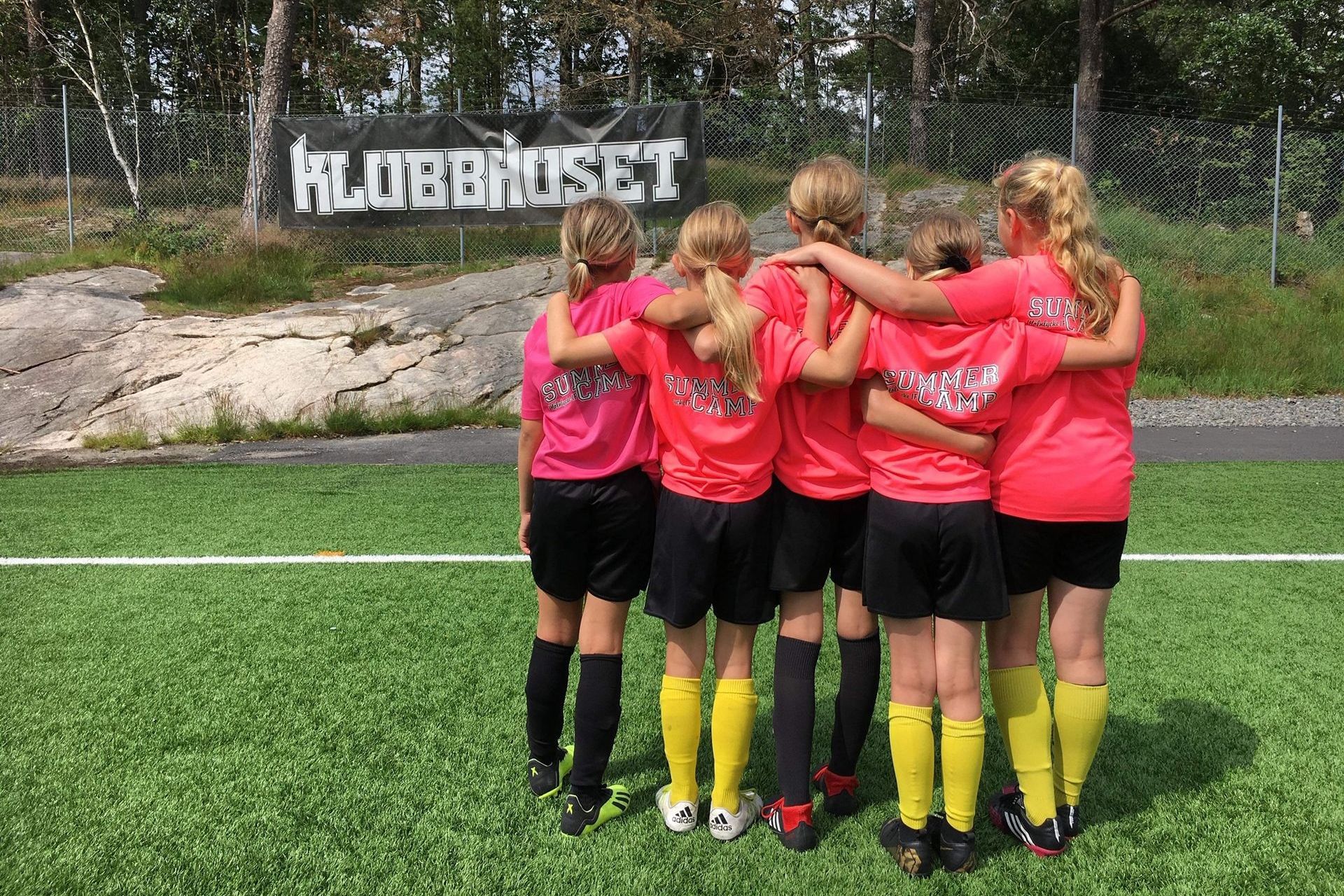 Flickor klädda i fotbollskläder, står på fotbollsplan och håller varandra om axlarna.