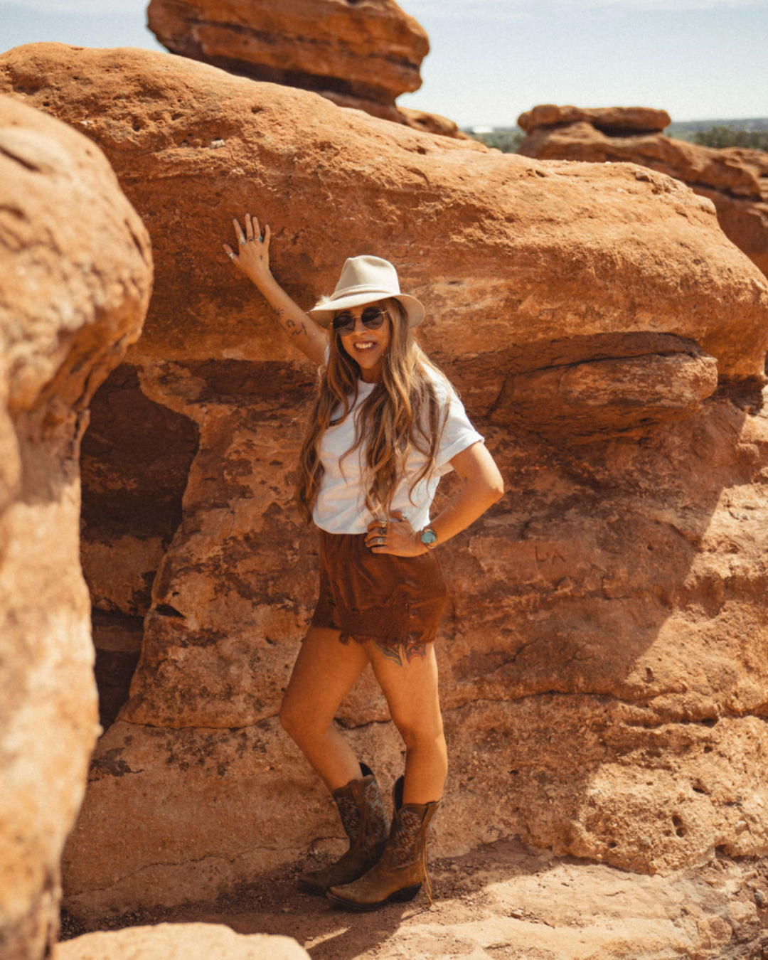 Woman in a white hat posing beside a red rock formation in a desert landscape.