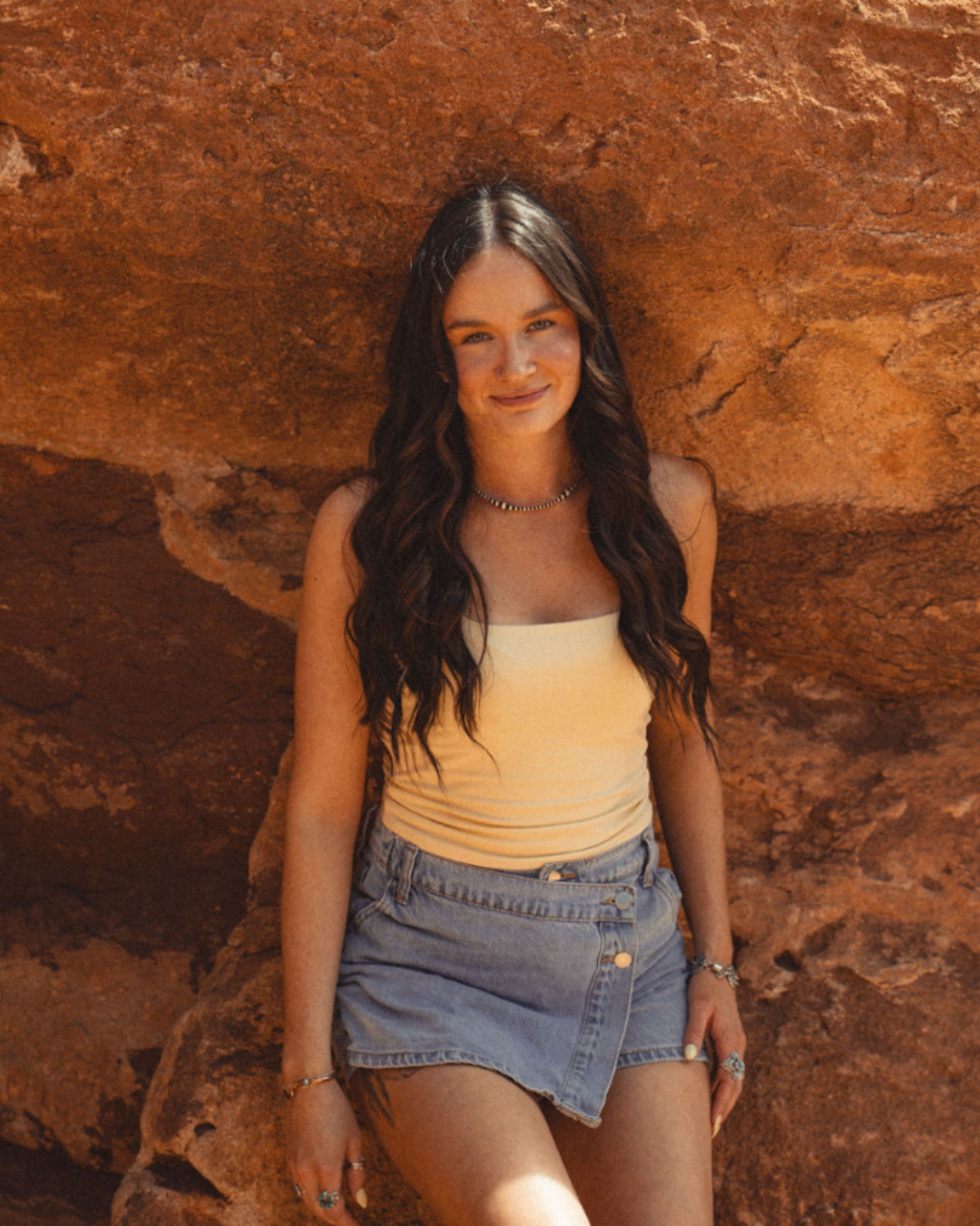 Woman standing against a rust-colored rock wall, wearing a cream top and denim shorts.