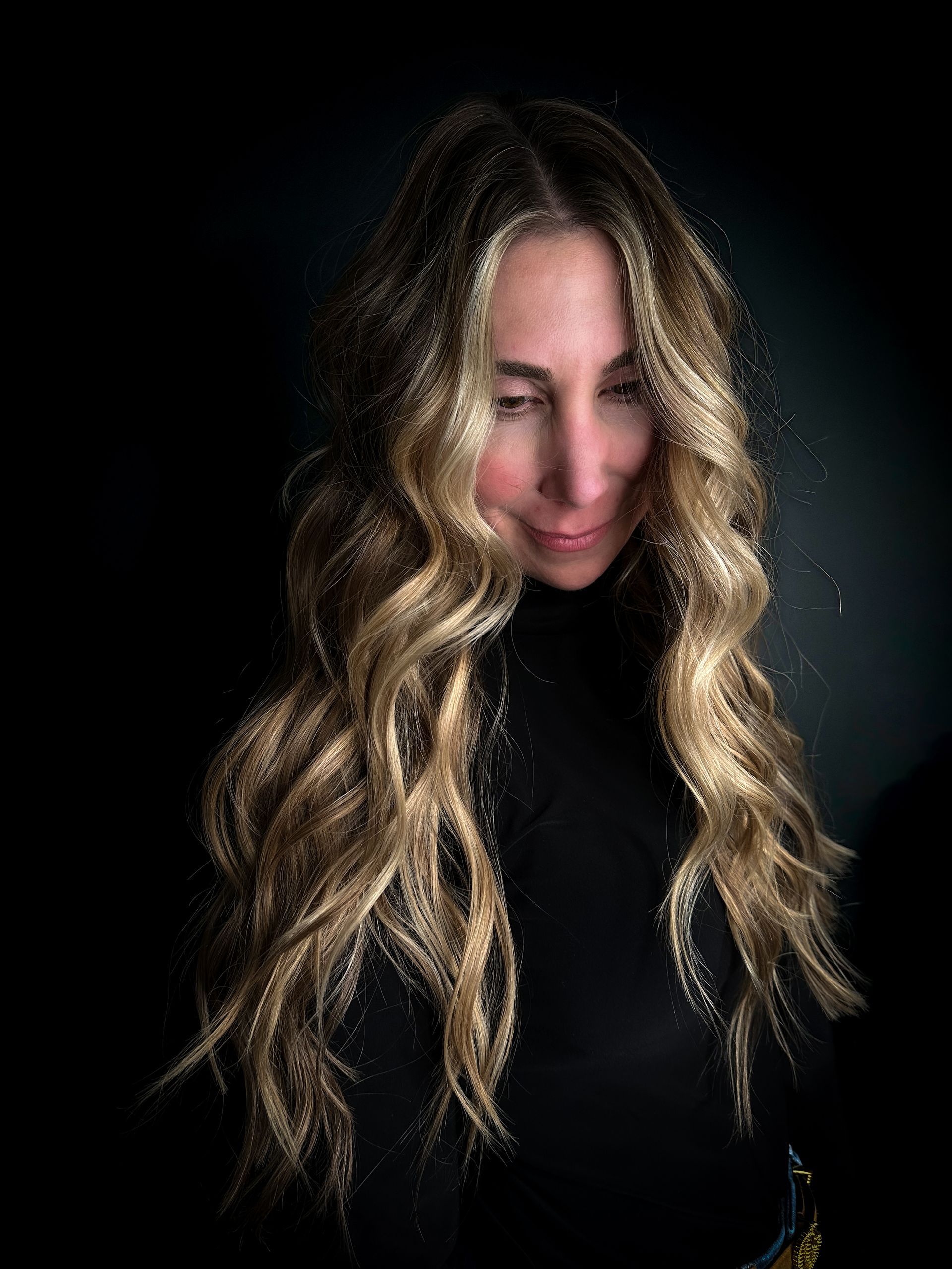 Woman with long wavy blonde hair in a black top against a dark background, looking down with a slight smile