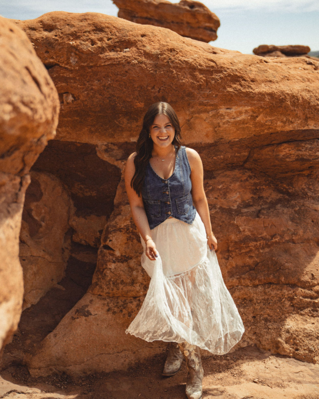 Woman in a blue top and white skirt standing by red rock formations in sunlight.