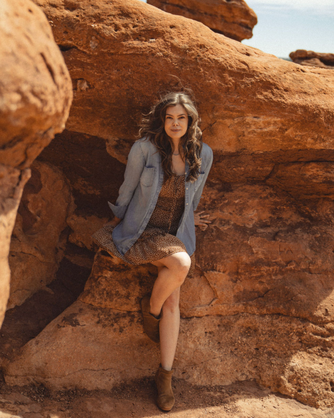 Woman sitting on red rock formations in a desert, wearing a gray jacket and patterned dress.