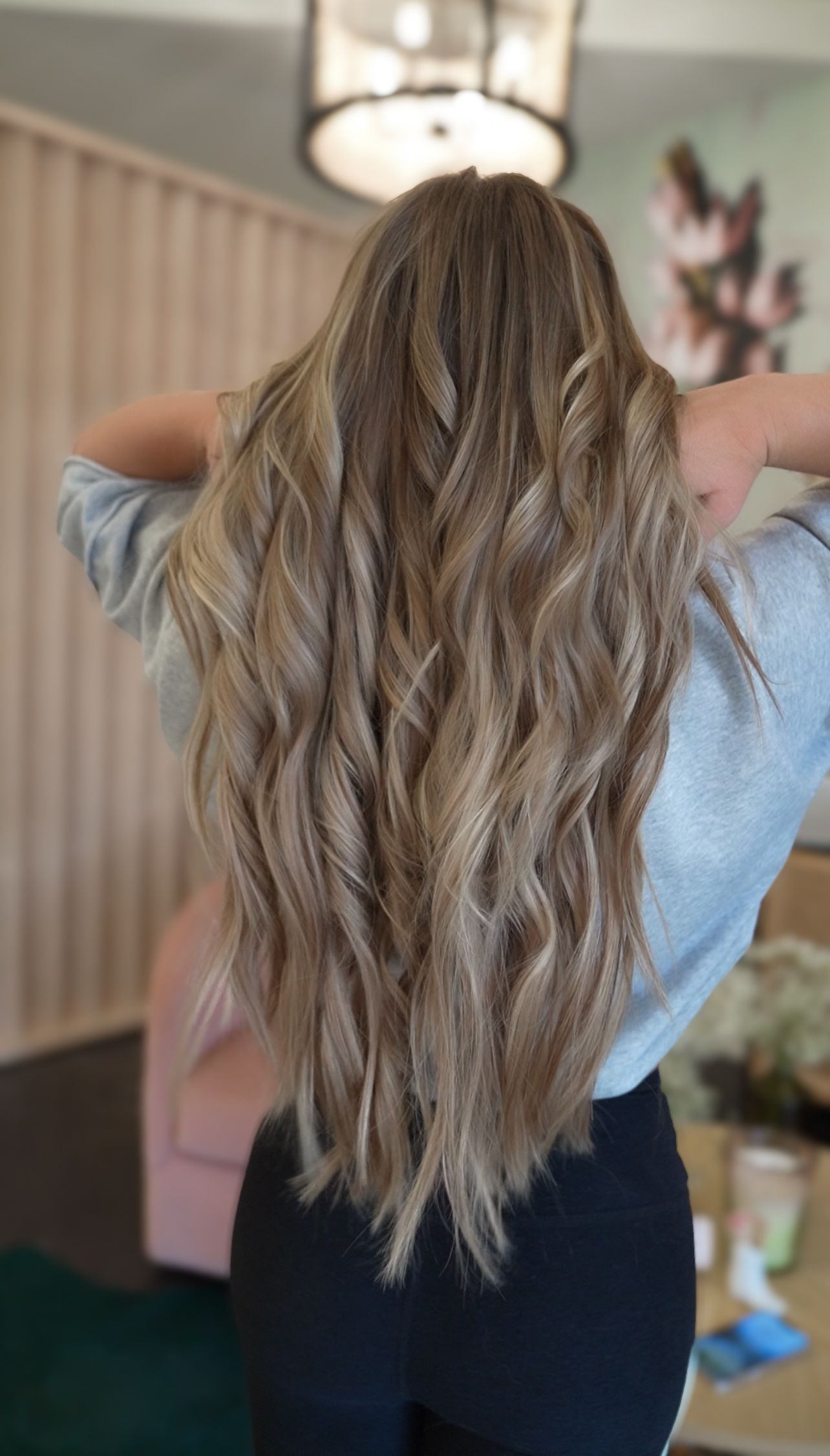 Person with long wavy blonde hair seen from behind in a room with a staircase and hanging light