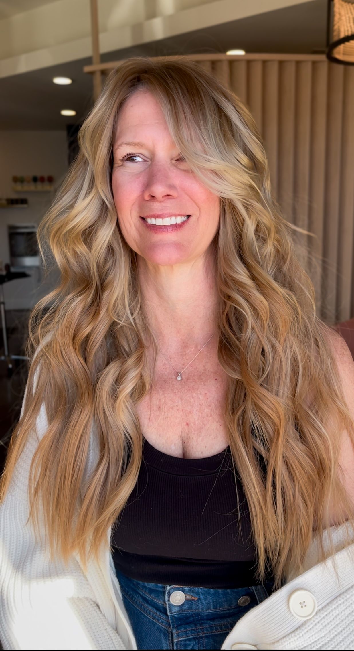 Smiling woman with long blonde hair in a black top, standing indoors near curtains.