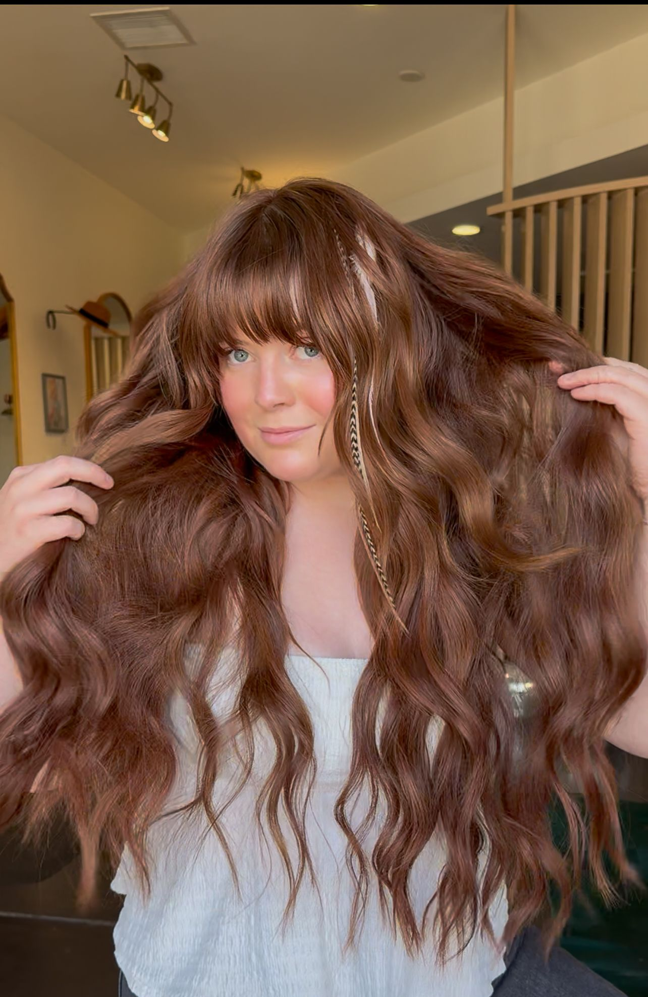 Person with long wavy brown hair holding it up indoors, wearing a white top.