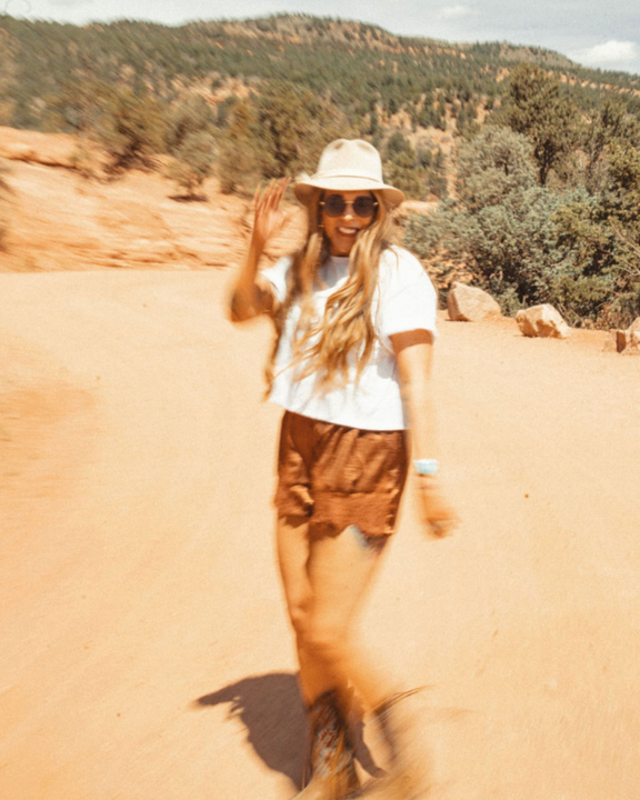 Woman in white hat and sunglasses walking on a sandy trail in a sunny desert landscape.