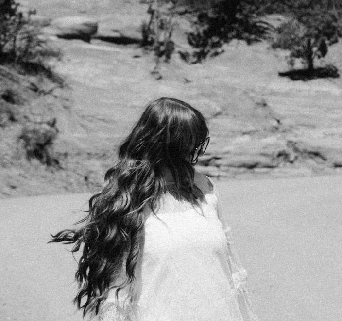 Long-haired person in a white dress standing by a riverbank in black and white.