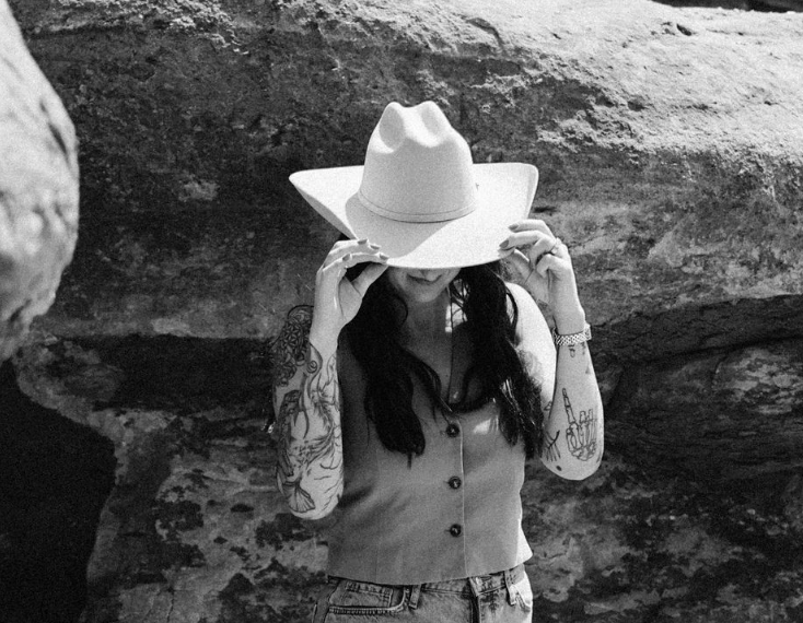 Woman in a cowboy hat adjusts it while standing against a rocky wall.