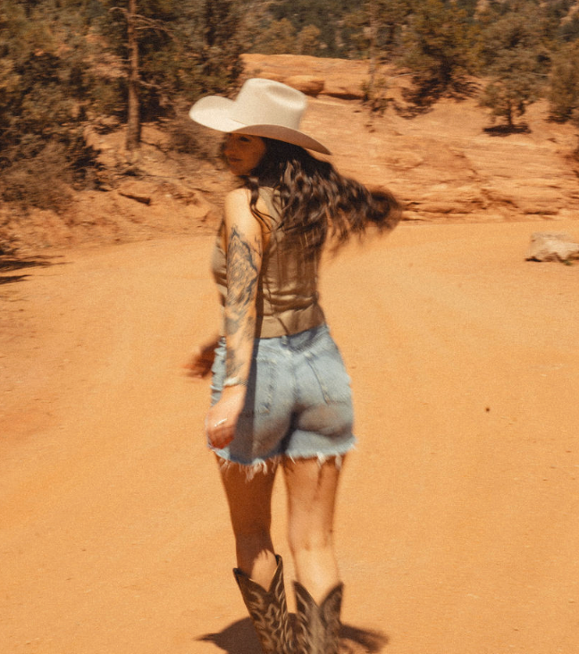 Person in a white cowboy hat walking on a sandy desert trail with a dog following behind
