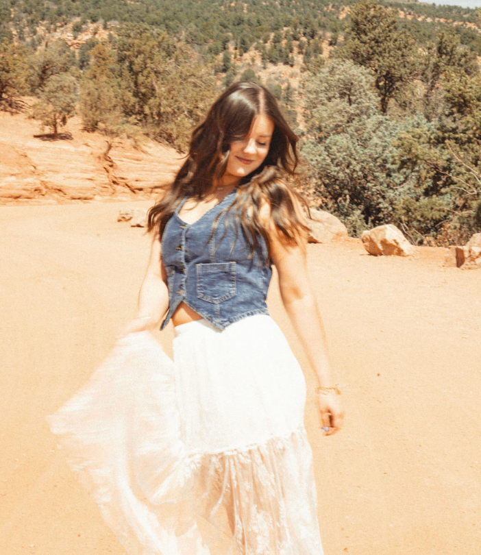 Woman in a blue top and white skirt standing in a sunny desert landscape with trees