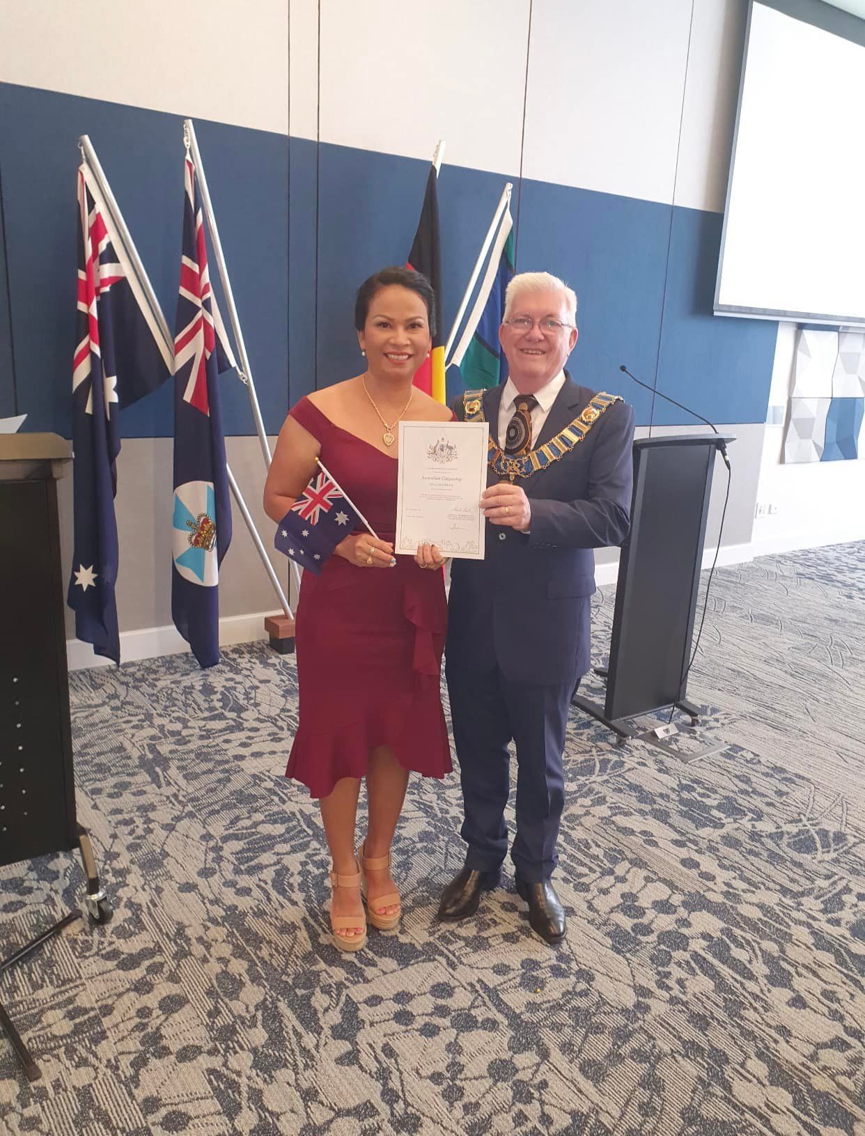 Woman in Red Dress Holding Certificate, Man in Suit, Flags, Room — Immigration Agents in Cairns North, QLD